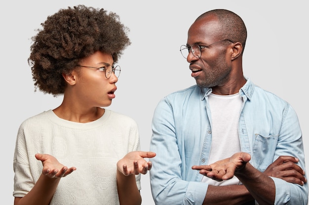 Casal afro-americano confuso se olha com raiva, aperta as mãos, não sabe onde o dinheiro sumiu, usa óculos redondos, isolados sobre uma parede branca. familia tem problemas