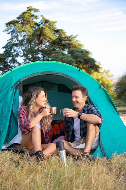 Casal alegre sentado na barraca, conversando e bebendo chá. Feliz caminhantes relaxando no gramado, acampando e curtindo a natureza. Viajantes ao ar livre na natureza. Conceito de turismo, aventura e férias de verão