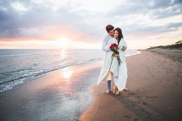 Casal andando na praia com um buquê de rosas ao pôr do sol