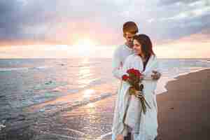 Foto grátis casal andando na praia com um buquê de rosas ao pôr do sol