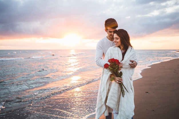 Casal andando na praia com um buquê de rosas ao pôr do sol