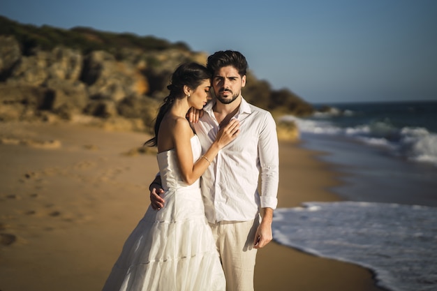 Casal apaixonado, caucasiano, vestindo roupas brancas e se abraçando na praia durante uma sessão de fotos de casamento