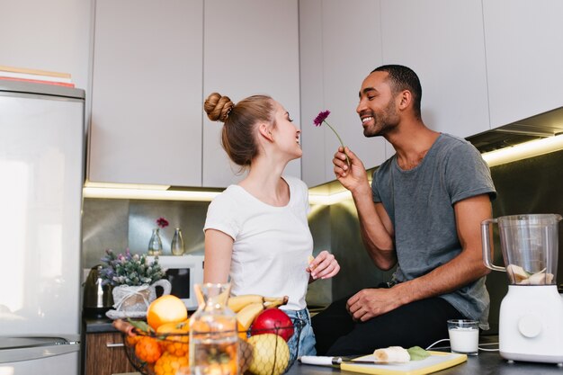 Casal apaixonado em camisetas flertando na cozinha. O marido dá uma linda flor à esposa. Rostos felizes, belo presente, alimentação saudável, par feliz.
