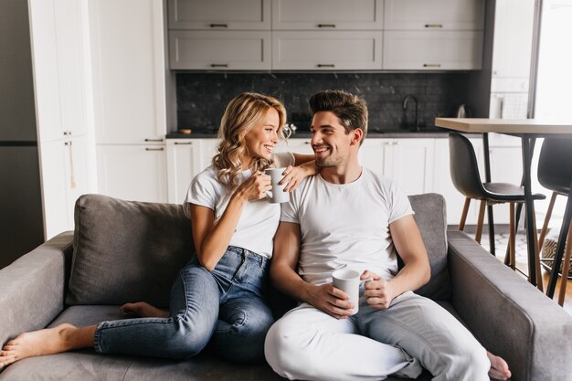 Casal apaixonado, sentado no sofá, segurando copos, olhando um para o outro e sorrindo. Casal romântico gosta de manhã juntos em casa.