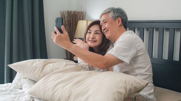 Casal asiático sênior selfie em casa. As avós, o marido e a esposa chineses sênior asiáticos felizes usando o selfie do telefone celular após acordam o encontro na cama no quarto em casa no conceito da manhã.