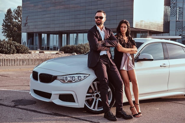 Casal atraente bem vestido, apoiando-se em um carro de luxo ao ar livre contra o arranha-céu.