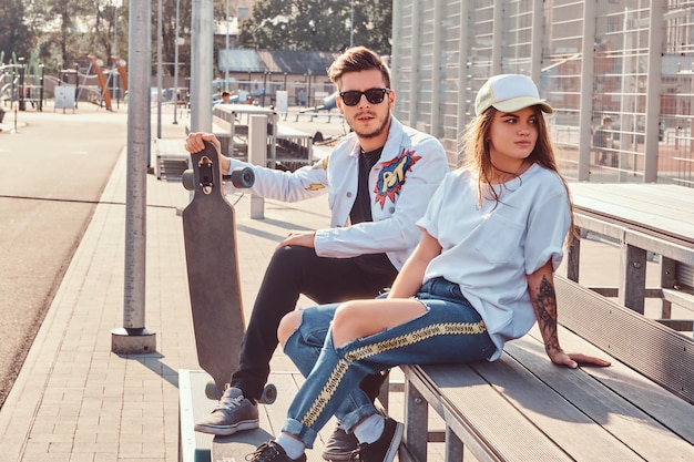 Casal atraente de jovens descolados vestidos na moda com skates sentados no banco no complexo esportivo da cidade em dia ensolarado, com tons quentes.