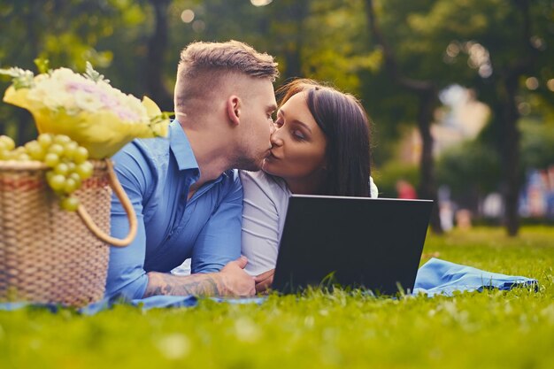 Casal atraente deitado em um cobertor em um gramado e está usando um laptop em um piquenique.