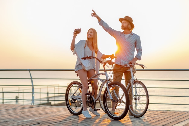 Casal atraente e feliz viajando no verão em bicicletas, homem e mulher com cabelo loiro boho hipster estilo moda se divertindo juntos, tirando fotos de passeios turísticos