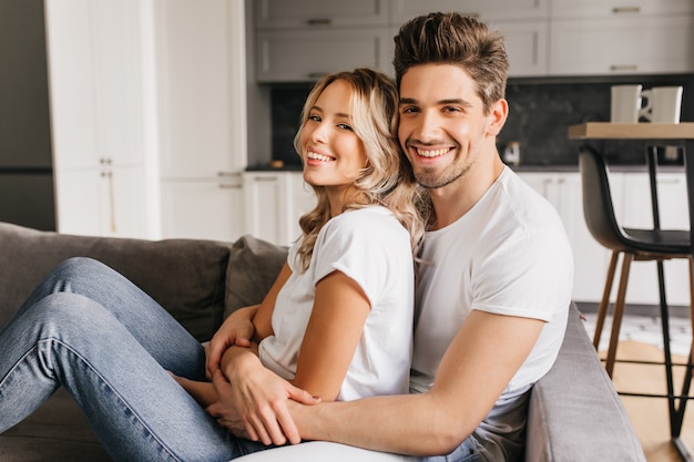 Casal atraente sorridente, sentado no sofá, abraçando um ao outro. Dois jovens felizes compartilham a manhã juntos.