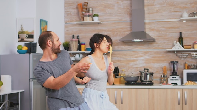 Casal autêntico dançando de pijama segurando utensílios de cozinha durante o café da manhã. Esposa e marido despreocupados rindo se divertindo, gozando a vida, autênticos casados, relação feliz positiva