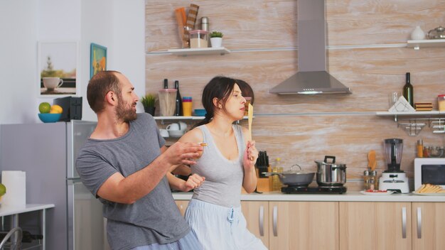 Casal autêntico dançando de pijama segurando utensílios de cozinha durante o café da manhã. Esposa e marido despreocupados rindo se divertindo, gozando a vida, autênticos casados, relação feliz positiva