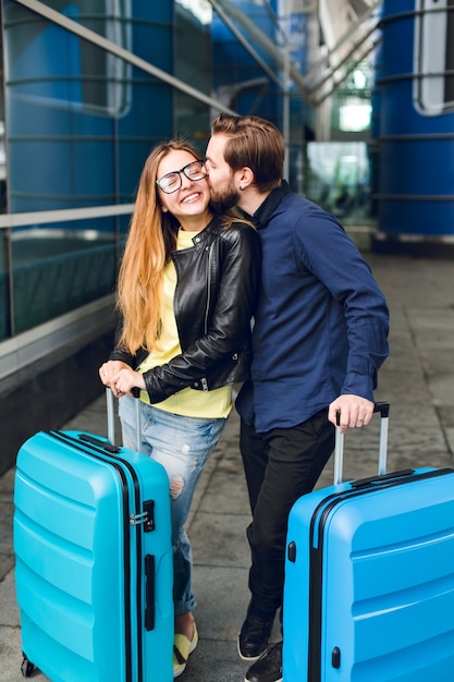 Casal bonito com malas está do lado de fora no aeroporto. ela tem cabelo comprido, óculos, suéter amarelo, jaqueta. ele usa camisa preta, barba. cara está abraçando e beijando a garota.