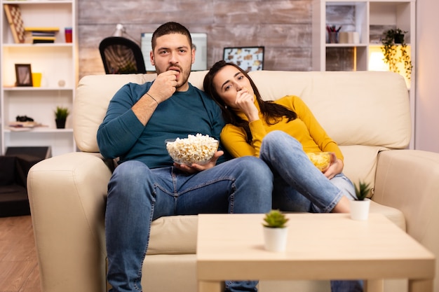 Casal bonito em casa comendo pipoca e assistindo tv no sofá da sala