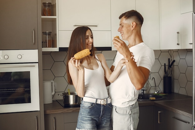 Casal bonito em uma cozinha. Senhora em uma camiseta branca. Os pares em casa comem milho cozido.