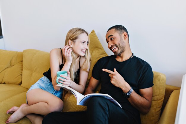 Casal brincando, conversando, se divertindo, sorrindo, sofá em casa. Os amantes relaxam, lêem uma revista, bebem chá. Humor positivo, provocação de conversa