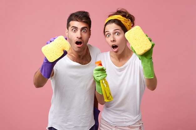 Casal chocado com esponjas e detergente para limpar vidros. Homem e mulher do serviço de limpeza vestidos casualmente, ficando surpresos ao ver muito trabalho. Conceito de pessoas, trabalho, limpeza e casa