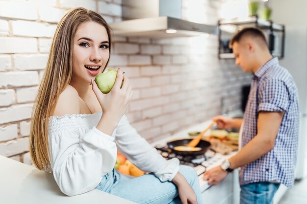 Casal cozinhando na cozinha. Mulher come a maçã.