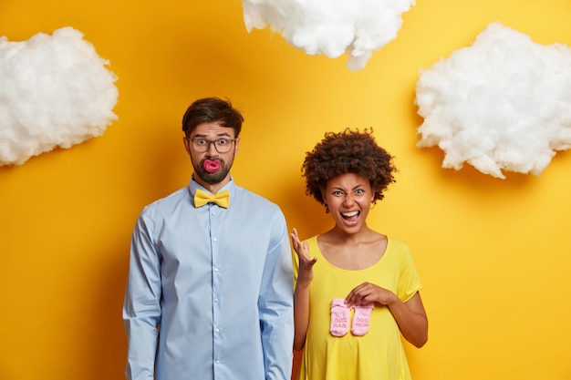 Casal da família se prepara para se tornarem pais posar com itens de bebê contra a parede amarela. Mulher afro-americana grávida emocional segura meias para o bebê sobre a barriga. Futuro pai barbudo chupando mamilo