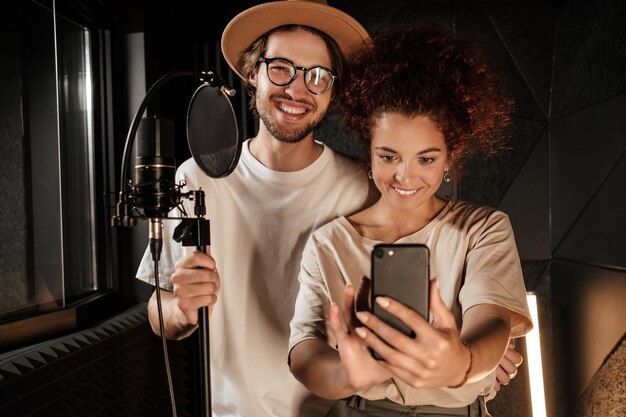 Casal de cantores elegantes atraentes alegremente tomando selfie juntos no smartphone no estúdio de gravação de som moderno