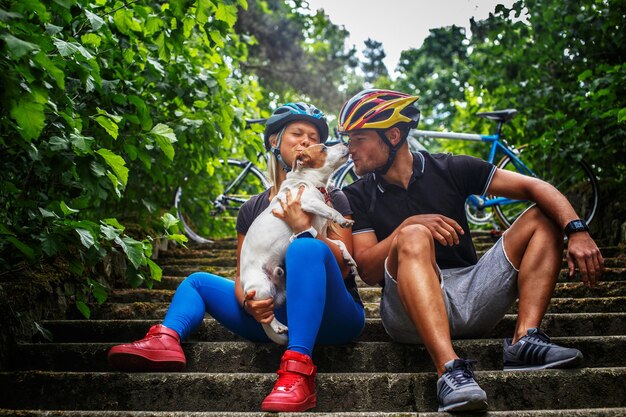 Casal de ciclistas com seu cachorro relaxa nos degraus em um parque primavera.