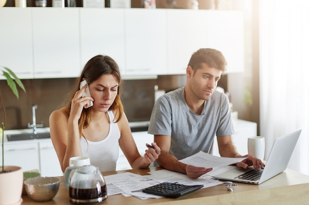 Foto grátis casal de família com problemas financeiros, tentando resolvê-los, vai tomar um empréstimo, ligar para o banco, assinar contrato. homem e mulher sentada na cozinha, usando dispositivos modernos para o seu trabalho de negócios