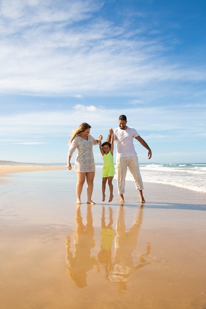 Casal de família feliz e menina curtindo caminhadas e atividades na praia, criança segurando as mãos dos pais, pulando e se pendurando