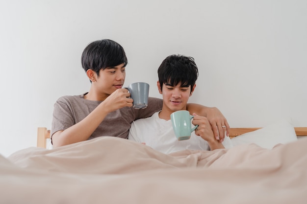 Casal de gays asiáticos falando divertindo-se em casa moderna. jovem amante ásia masculino feliz relaxar resto beber café depois de acordar enquanto estava deitado na cama no quarto em casa de manhã.