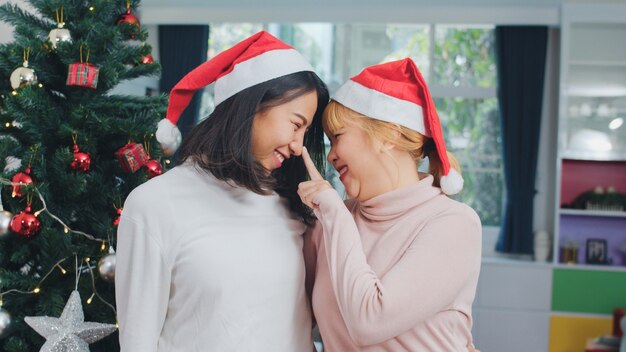 Casal de lésbicas asiáticas comemorar o festival de Natal. LGBTQ feminino adolescente usar chapéu de Natal relaxar feliz sorrindo olhando desfrutar de férias de inverno xmas juntos na sala de estar em casa.