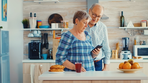 Foto grátis casal de mais velhos durante o bate-papo por vídeo com a família usando o smartphone na cozinha. conversa online dos avós. idosos com tecnologia moderna em idade de aposentadoria usando aplicativos móveis