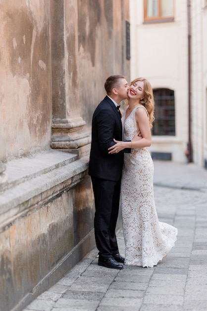 Casal de noivos está beijando ao ar livre perto da parede, feliz casal sorriu, loucamente apaixonado