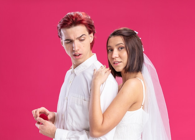Casal de noivos felizes posando juntos, homem segurando uma caixa vermelha com um anel de casamento e sua noiva vestida de branco em pé atrás da parede rosa