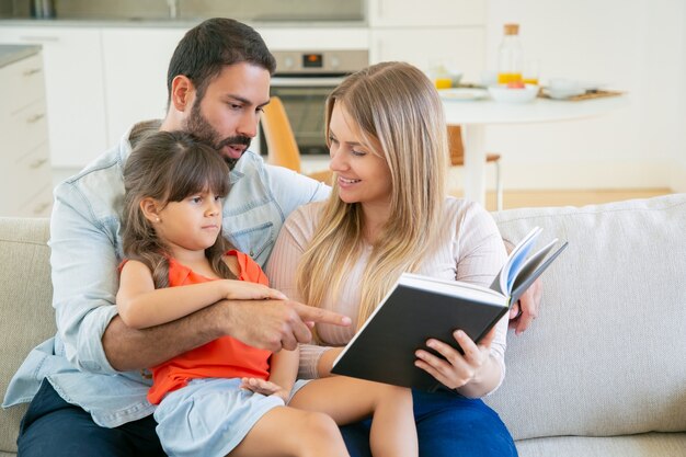 Casal de pais felizes e a garotinha de cabelo preto, sentado no sofá na sala de estar e lendo o livro juntos.