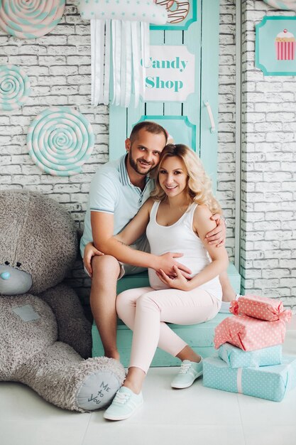 Casal de positividade de homem bonito e mulher grávida bonita loira sentada na escada no estúdio abraçando o estômago e gesticulando o coração pelas mãos Jovem família elegante no interior de hortelã doce entre brinquedos