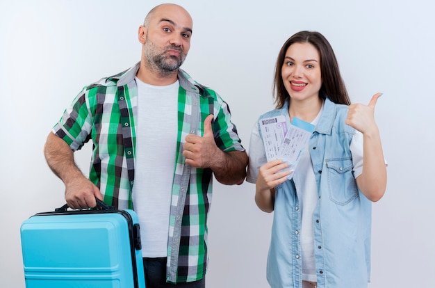 Casal de viajantes adultos confiante segurando uma mala e uma mulher sorridente segurando passagens, ambos mostrando o polegar para cima, olhando