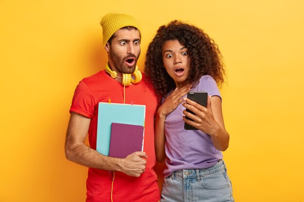 Casal elegante chocado posando contra a parede amarela com gadgets