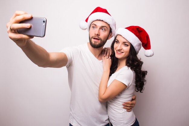 Casal feliz com chapéu de Papai Noel acenando e tirando selfie no smartphone, isolado no fundo branco