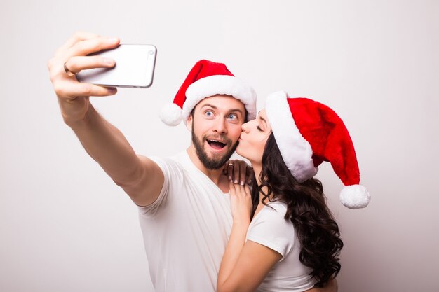 Casal feliz com chapéu de Papai Noel acenando e tirando selfie no smartphone, isolado no fundo branco