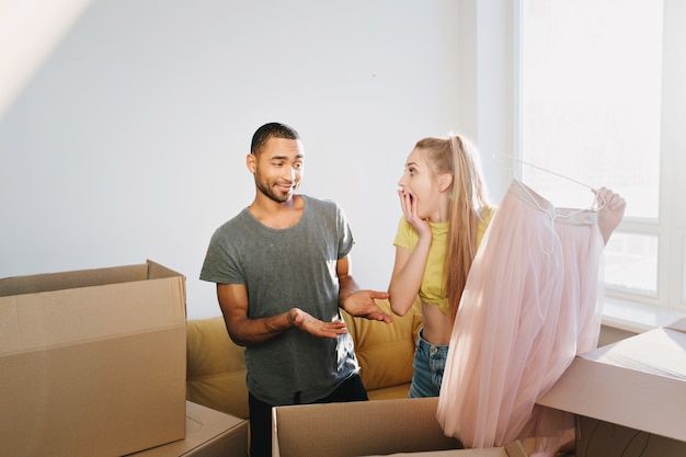 Foto grátis casal feliz comprou a casa, a família mudou-se para um novo apartamento, inauguração de casa, desempacotando caixas. mulher encontrada presente, marido surpreendido esposa, saia rosa de presente. homem vestindo camiseta cinza, top feminino amarelo.