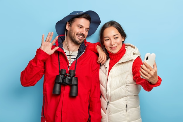 Casal feliz de raça mista tira uma selfie no smartphone, aproveita a jornada de caminhada, fica perto um do outro, vestido com roupa casual, usa binóculos