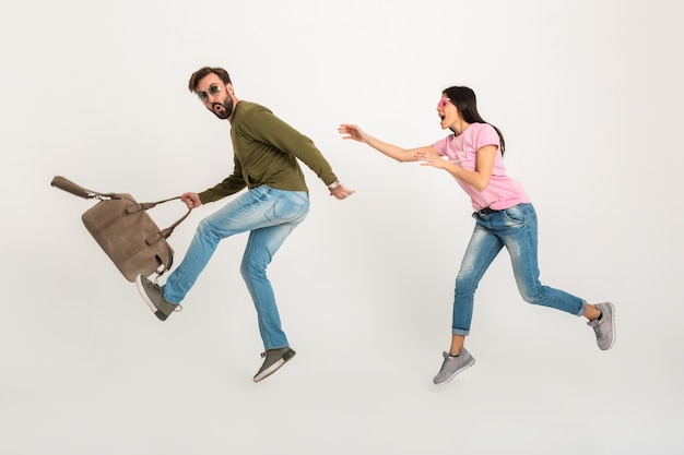 Casal feliz e engraçado pulando isolado, mulher muito sorridente em uma camiseta rosa correndo atrás de um homem de moletom segurando uma bolsa de viagem, vestido de jeans