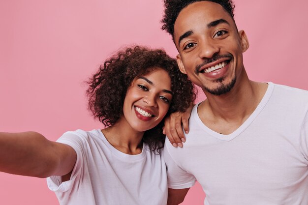 Casal feliz em camisetas brancas tirando uma selfie na parede rosa