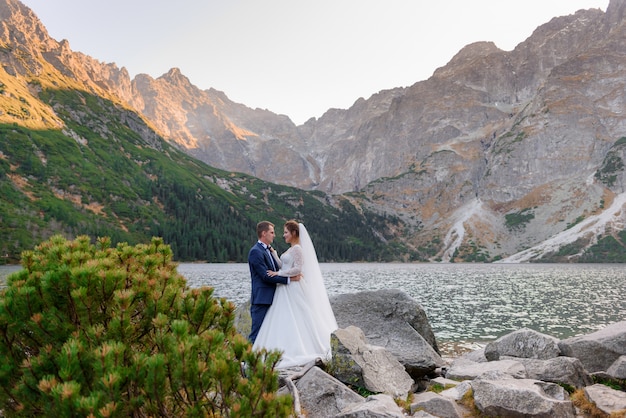 Casal feliz no amor, vestido com roupas de casamento está quase se beijando com uma vista deslumbrante das montanhas e do lago das montanhas