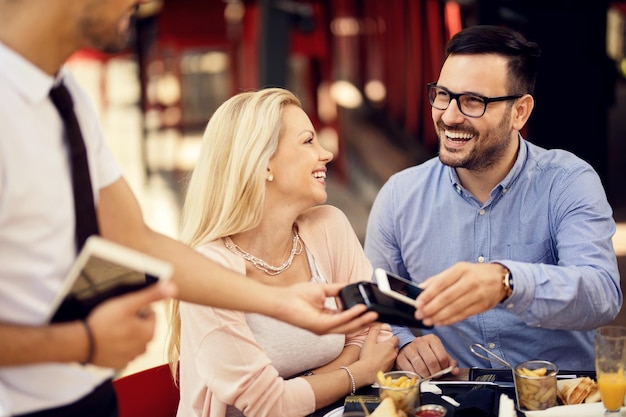 Casal feliz pagando conta depois do almoço em um bar o homem está usando telefone inteligente para pagamento sem contato