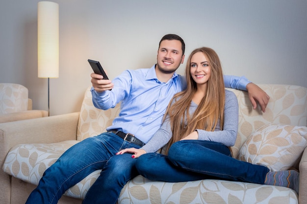 Casal feliz sentado no sofá com controle remoto nas mãos e assistindo televisão juntos