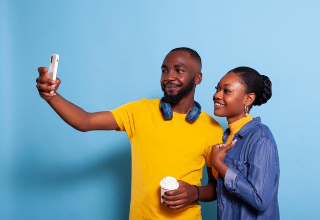 Casal feliz tirando selfies no smartphone em estúdio, fazendo memórias juntos. Namorado e namorada modernos tirando fotos no celular, abraçando e usando a tecnologia sobre o fundo.