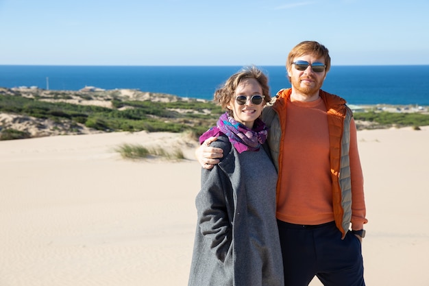 Casal feliz vestindo roupas quentes, passando o tempo de lazer no mar, em pé na areia, se abraçando, olhando para a frente