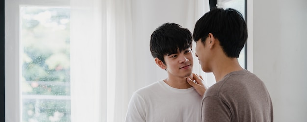 Casal gay asiático em pé e abraçando perto da janela em casa. Os jovens asiáticos LGBTQ + homens se beijando felizes relaxam o resto juntos passam um tempo romântico na sala de estar da casa moderna de manhã.