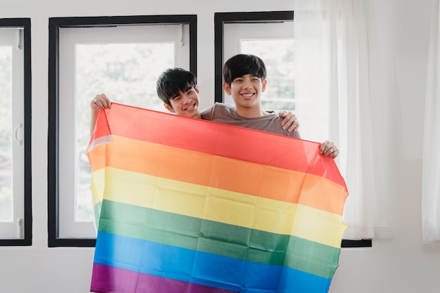 Casal gay asiático novo do retrato que sente feliz mostrando a bandeira do arco-íris em casa. os homens da ásia lgbtq + relaxam o sorriso, olhando para a câmera enquanto abraçam na moderna sala de estar em casa de manhã.