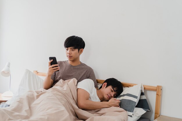 Casal gay asiático usando telefone celular em casa. Jovem Ásia LGBTQ + homem feliz relaxar resto depois de acordar, verifique as mídias sociais enquanto seu namorado dorme deitado na cama no quarto em casa de manhã.